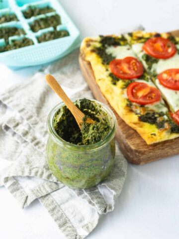 Basil pesto in a short jar with a small wooden spoon with pizza in the background