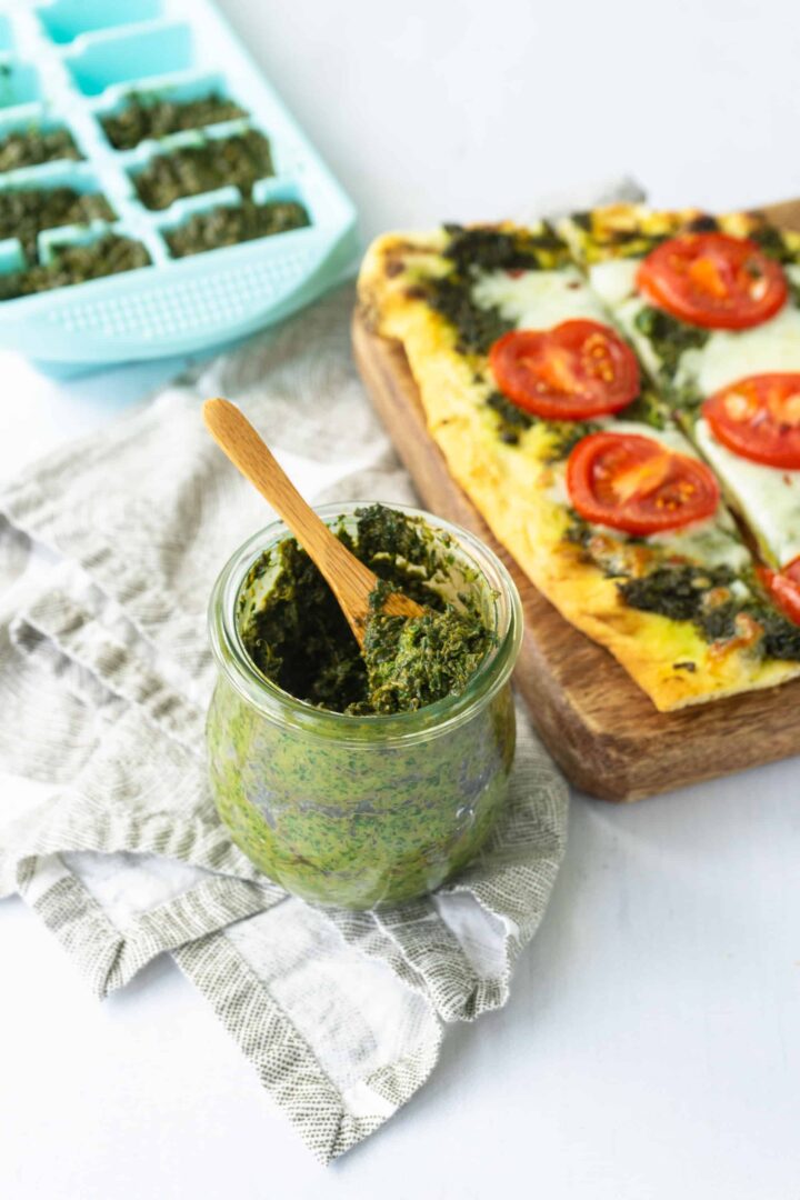 Basil pesto in a short jar with a small wooden spoon with pizza in the background