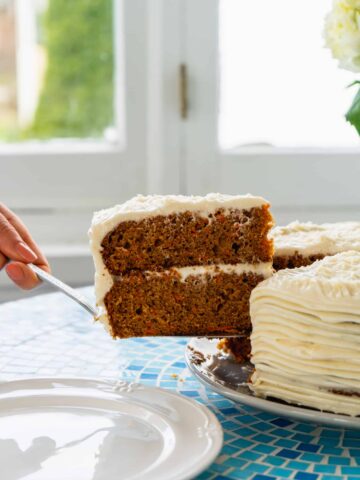 Slice of carrot cake with cream cheese frosting being removed from whole cake on blue table