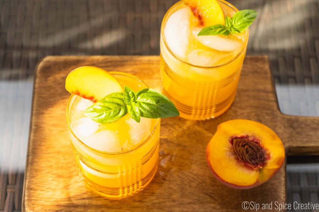 Basil Peach Whiskey Cocktails in the late summer light with a half a peach on a wooden board